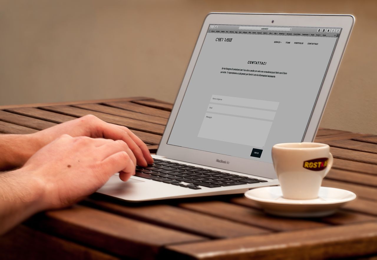 Gros plan sur des mains tapant sur un ordinateur portable, à côté d’une tasse de café posée sur un bureau en bois.